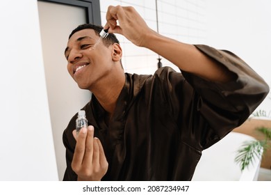 Black Man Wearing Bathrobe Applying Face Serum At Morning In Bathroom