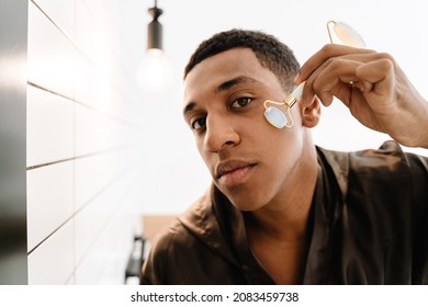 Black Man Wearing Bathrobe Massaging Face With Jade Roller In Bathroom