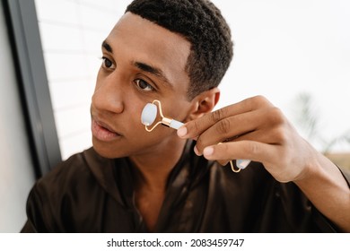 Black Man Wearing Bathrobe Massaging Face With Jade Roller In Bathroom