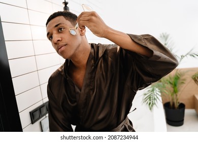 Black Man Wearing Bathrobe Massaging Face With Jade Roller In Bathroom