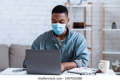 Black Man Wearing Face Mask Working On Laptop Sitting In Modern Office. Social Distancing At Work During Coronavirus Covid-19 Pandemic. Remote Work Concept. Selective Focus