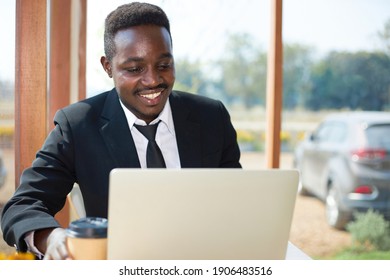 A Black Man Wearing Black Suit Sitting Inside A Cafe With A Laptop, Sat By A Glass Window With A Coffee Cup.