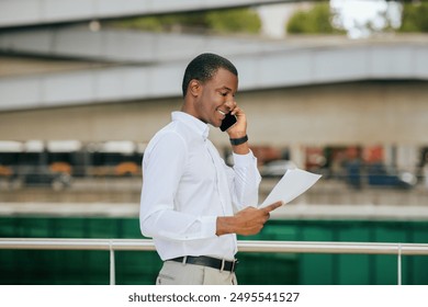 Black man wearing a white long-sleeved shirt is talking on his phone while reviewing documents. He is standing outside in front of a modern building with a green railing. - Powered by Shutterstock