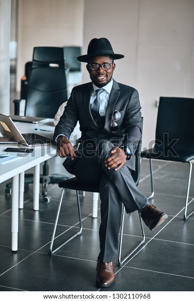 black man\
wears fashion hat, suit sitting on the office chair and looking at\
the camera . full length photo. free\
time