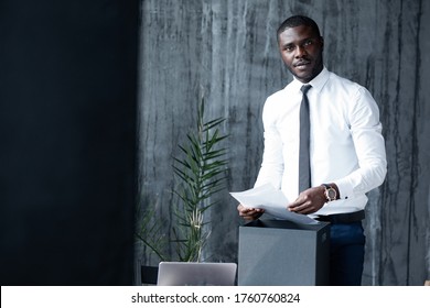 Black Man In A White Shirt Standing Before A Desk And Holding A Document And Looking At The Camera.