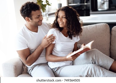 Black Man And Woman Are Sitting On The Couch. A Woman Is Reading A Book, A Man Is Sitting And Hugging Her. They Smile. They Rest At Home.