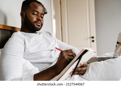 Black Man Writing Down Notes And Working With Laptop While Lying In Bed At Home