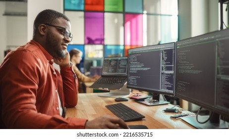 Black Man Writing Lines Of Code On Desktop PC With Two Monitors And A Laptop In Stylish Office. Professional Male Developer Programming Artificial Intelligence Software For Start-Up Company.