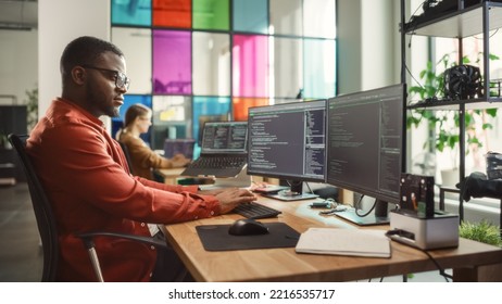 Black Man Writing Lines Of Code On Desktop PC With Two Monitors And A Laptop Aside In Stylish Office. Professional Male Developer Programming Artificial Intelligence Software For Start-Up Company.