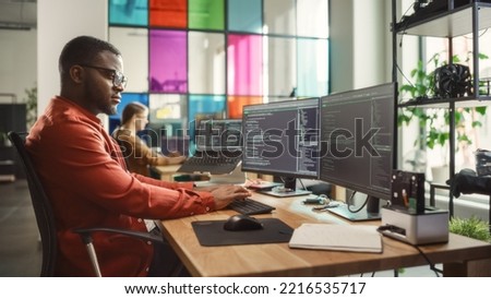 Black Man Writing Lines of Code On Desktop PC With Two Monitors and a Laptop Aside in Stylish Office. Professional Male Developer Programming Artificial Intelligence Software for Start-Up Company.