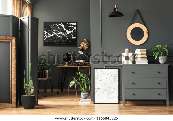 Black map on grey wall in dark living\
room interior with plants and poster. Real\
photo