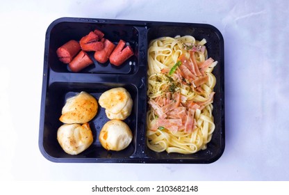 A Black Meal Box Consist Of Carbonara Spaghetti, Cheese Bread Balls And Beef Sausages On White Background, High Angle View