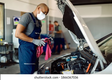 Black mechanic wearing face mask while repairing car's engine under the hood in a workshop.  - Powered by Shutterstock