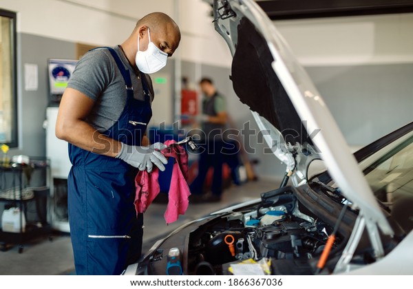 Black mechanic wearing face mask\
while repairing car\'s engine under the hood in a workshop.\
