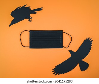 Black Medical Mask And Paper Crows With A Witch Lie Diagonally On An Orange Background With Space For Text, Flat Lay Close-up. Scary Concept And Coronavirus.lack Mask And Paper Crow On Orange.