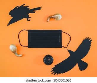 Black Medical Mask, Three Decorative Pumpkins And Paper Crows With A Witch Lie On An Orange Background With Space For Text, Close-up. Scary Concept And Coronavirus.