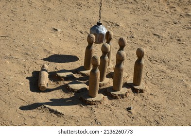 black metal chairs and round tables in a cafe on a cobbled square with a sand surface of compacted yellow gravel under an old tree. Russian wooden skittles and balls on a chain