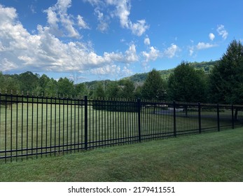 A black metal privacy fence runs in front of a rural property with a field and trees in the summer in America