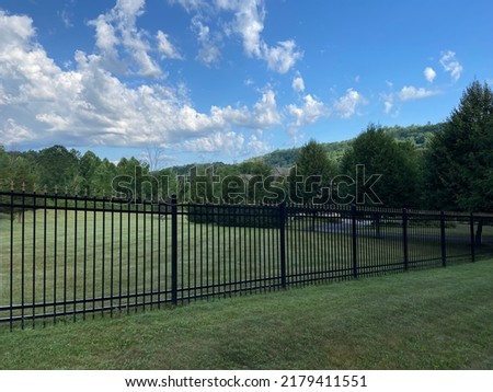 A black metal privacy fence runs in front of a rural property with a field and trees in the summer in America