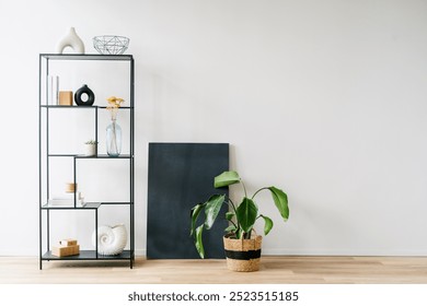 Black metal shelving unit filled with decorative objects sits against a minimalist copy space wall. Potted plant and abstract painting on the wooden floor, creating serene atmosphere in apartment - Powered by Shutterstock