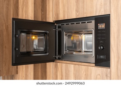 A black microwave with open door set into a wooden kitchen cabinet. Empty turntable glass plate for food inside oven, side view