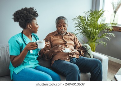 A black mid-adult female nurse in teal scrubs attentively provides care to an elderly black male patient, seated comfortably on a couch in a home setting. - Powered by Shutterstock