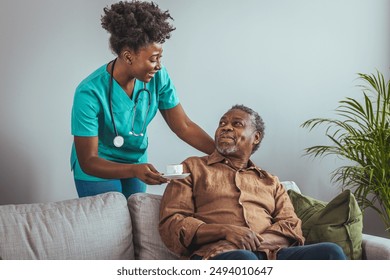 A black mid-adult nurse, in scrubs and with a stethoscope, attentively checks the temperature of a senior black male comfortably seated at home. - Powered by Shutterstock