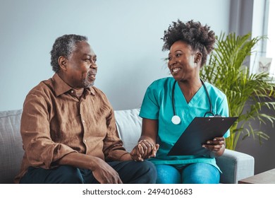 A black mid-adult nurse takes notes while engaging with an elderly male patient in a home setting, providing medical care and attention with a smile. - Powered by Shutterstock