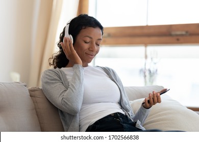 Black Middle Aged Woman Listen Music With Her Eyes Closed. Calm Home Weekend, Relax While Sit On Sofa, Enjoy Favorite Tune, Popular Music, Use Modern Wireless Headphones And Gadgets In Living Room