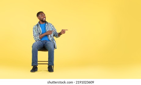 Black Millennial Man Pointing Fingers Aside At Free Space Sitting In Chair Over Yellow Studio Background. Male Advertising Offer Showing Your Text. Panorama, Full Length