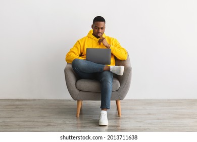 Black Millennial Man Using Laptop Thinking Writing Article For Blog, Browsing Internet Sitting In Armchair Near Gray Wall At Home. African American Male Freelancer Working Online On Computer