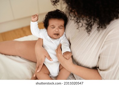 Black millennial mom calms crying sad little baby on white bed in bedroom. Disease treatment, teeth growing, child care, parenthood and family at home, health care and love - Shutterstock ID 2311020547