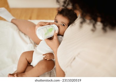 Black millennial mother feeds from bottle hungry little baby on white bed, enjoys love and comfort in bedroom. Kid food with vitamins, baby care, parenthood and family at home - Shutterstock ID 2311020519