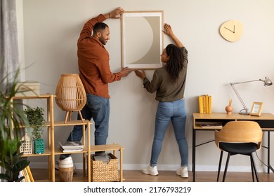 Black Millennial Spouses Hanging Poster In Frame On Wall Together Standing At Home. Married Couple Decorating Living Room After Renovation In New House Indoors. Interior Decoration And Design