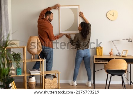 Black Millennial Spouses Hanging Poster In Frame On Wall Together Standing At Home. Married Couple Decorating Living Room After Renovation In New House Indoors. Interior Decoration And Design Foto stock © 