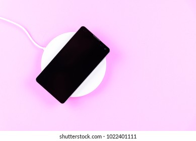 Black mobile phone put on white wireless charger in pink pastel background