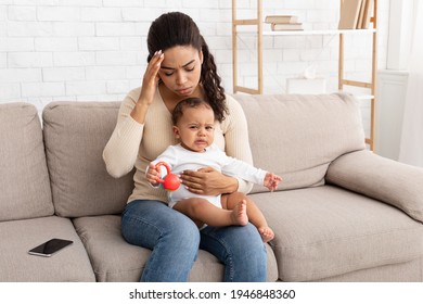 Black Mom With Baby Having Headache Suffering From Migraine Pain Sitting With Crying Toddler Son At Home. Sick Mom Touching Aching Head Caring For Little Infant Indoors. Healthcare Concept