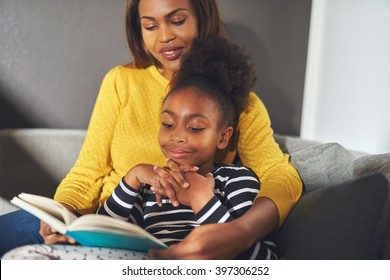 Black Mom And Daughter Reading A Book Sitting On Sofa Smiling