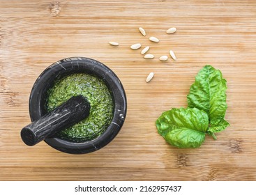 Black mortar full of basil pesto, pine nuts ans basil leaves on wooden table, top view of italian pesto recipe for pasta