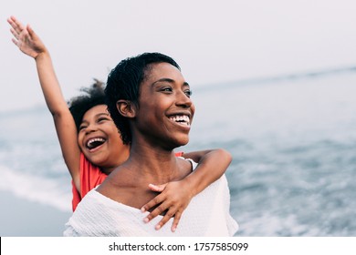 Schwarze Mutter und Tochter, die im Sommerurlaub bei Sonnenuntergang am Strand rennen - Familienmitglieder, die sich im Freien amüsieren - Reisen und fröhliches Leben - Fokus auf das Gesicht der Mutter