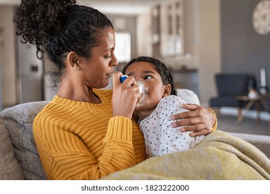 Black Mother Helping Sick Daughter Use Nebulizer While Embracing Her On Couch At Home. Woman Makes Inhalation With Equipment To Indian Girl. Ill Child Having Respiratory Illness Helped By Lovely Mom.