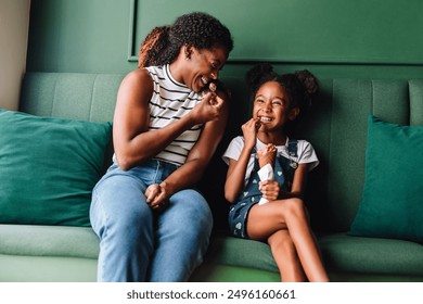 A black mother and her daughter laugh together while smiling and eating ice cream, sitting on a green sofa. - Powered by Shutterstock