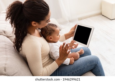 Black Mother With Toddler Baby Video Calling Using Tablet Waving Hand To Empty Computer Screen Sitting On Sofa Indoor. High Angle, Side View, Mockup. Mom And Infant Boy Calling Dad