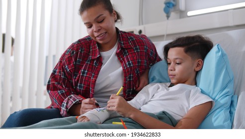 Black Mother Visiting Son Lying With Broken Arm In Bed In Hospital Ward. Happy African Kid Painting Arm Plaster Cast With Mom Having Fun Together In Clinic Room