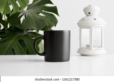 Black Mug Mockup With A Candle Holder On A White Table And A Monstera Plant.