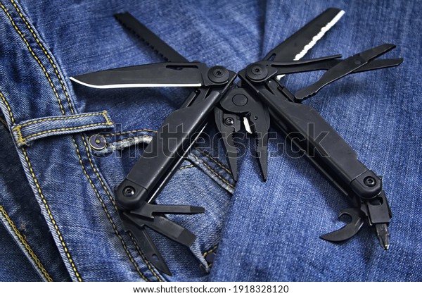 Black\
multi tool  ( multitool ) with expanded tools and pliers close up\
on denim backround. Business and craft\
concept.