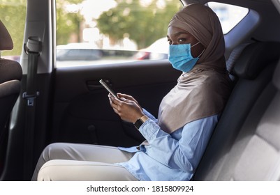 Black muslim businesswoman wearing face mask while using taxi during coronavirus pandemic, sitting on backseat of car and browsing smartphone, having personal driver, going to office, side view - Powered by Shutterstock