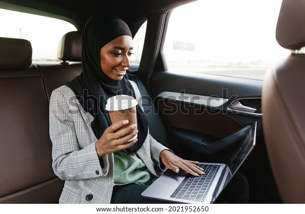 Black muslim woman wearing headscarf\
working with laptop and drinking coffee in\
car