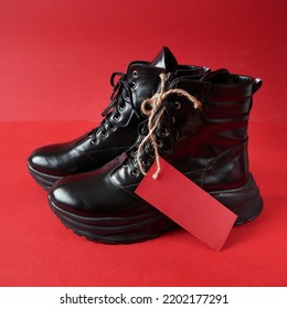 Black New Woman Or Man Rough Pair Of Boots With A Tag For The Inscription From The Sale On A Red Background, Autumn And Winter Sale Concept