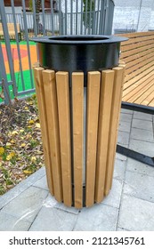 A black outdoor trash can with wooden trim stands next to a bench, close-up.